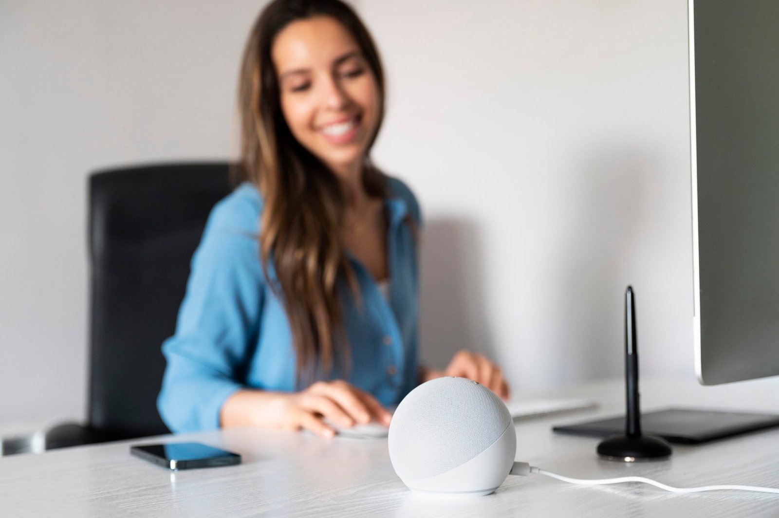 Mulher sentada em frente a computador. Sobre a mesa, uma assistente virtual do tipo Alexa